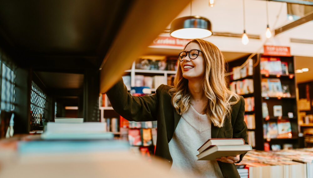 Photo of young blonde woman with glasses looking at book shelves at bookstore. Young woman is looking for new books to buy. Photo of young blonde woman with glasses looking at book shelves at bookstore. Young woman is looking for new books to buy.